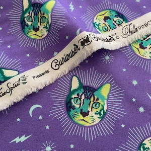 May include: Purple fabric with a repeating pattern of cat faces in teal, yellow, and black, surrounded by white sunbursts. The fabric also features stars, moons, and lightning bolts. A white fabric strip with text that reads "Free Spirit Presents Curiouser by Tula Pink" is diagonally across the image.