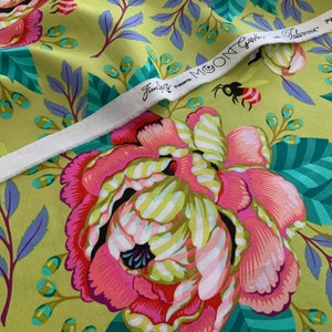 Op de afbeelding: Een limoengroene stof met een groot bloemenpatroon van roze en witte pioenen, groenblauwe bladeren en kleine bijen. Een witte zelfkantstrook loopt diagonaal over het oppervlak, bedrukt met de tekst FreeSpirit presents MOON GARDEN by Tula Pink PWTP 195.