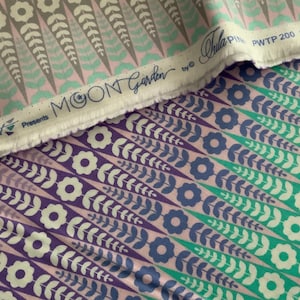 Op de afbeelding: Een stuk stof met een herhalend patroon van gestileerde bloemen en bladeren in tinten paars, groenblauw en roze. De witte zelfkant toont de tekst Free Spirit Presents MOON Garden by Tula Pink PWTP 200 Oopsie Daisy.
