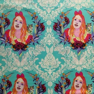 Op de afbeelding: Een turquoise stof met een herhalend patroon van een persoon met lang oranje haar en een roze strik, met een parelketting en een rode jurk. De persoon is omringd door een bloemenkrans en een kleine kat, tegen een witte damast achtergrond.