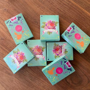 May include: Several decks of playing cards with teal boxes. Each box features a large rose with a banner and the words "Enchant & Charm". The top of the boxes have a pink circle with a silhouette design. The cards are arranged on a wooden surface.