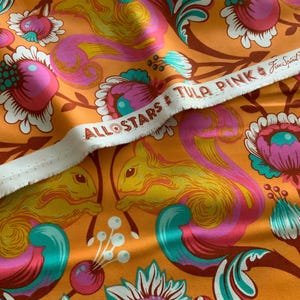 Op de afbeelding: Een levendige oranje stof met een herhalend patroon van gele eekhoorns, roze en turquoise gestileerde bloemen en bruine stelen. Een witte zelfkant loopt diagonaal over de print met de tekst ALL STARS TULA PINK Free Spirit.