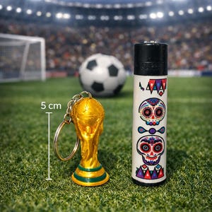 May include: A gold-colored trophy keychain and a white lighter with Day of the Dead skull designs are displayed on a green field. A soccer ball is in the background. The lighter is decorated with colorful skull illustrations.