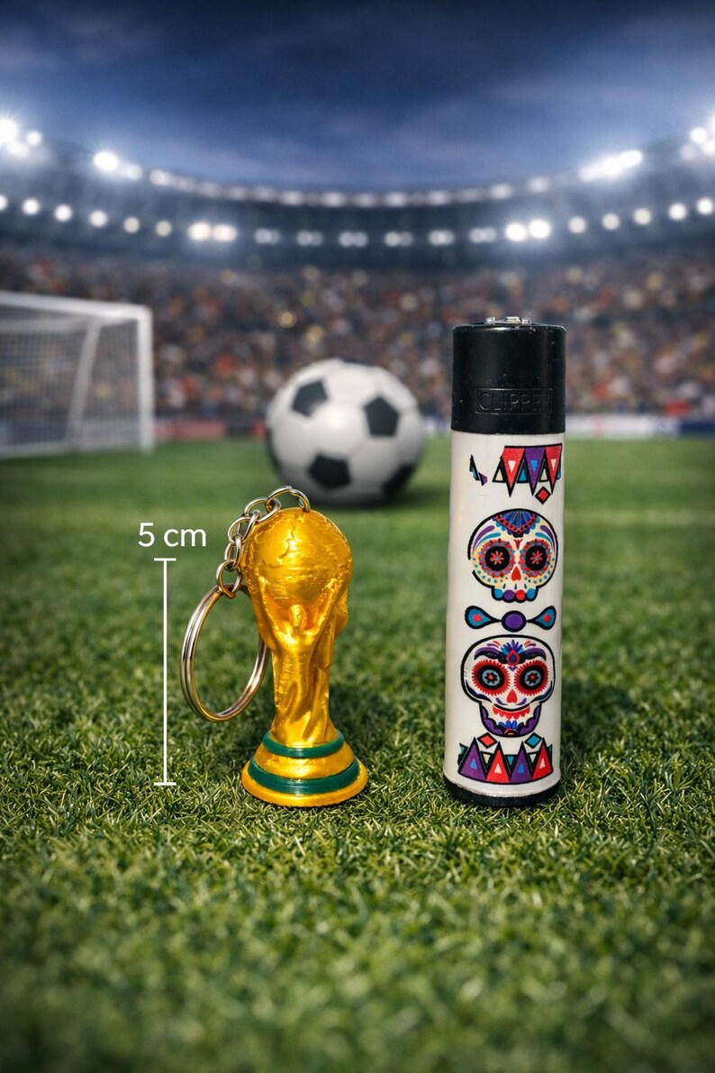May include: A gold-colored trophy keychain and a white lighter with Day of the Dead skull designs are displayed on a green field. A soccer ball is in the background. The lighter is decorated with colorful skull illustrations.