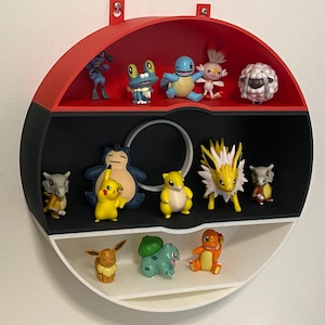 May include: A circular shelf unit in red, black, and white, designed like a Poké Ball, filled with Pokémon figurines. The shelf has three tiers, each showcasing different characters in bright colours. The unit is mounted on a white wall.