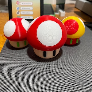 May include: Three mushroom-shaped figurines in red, white, and yellow. The red mushrooms have white spots and green bases, while the yellow mushroom has a red center. The figurines are on a gray surface.