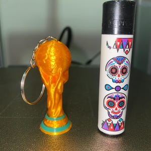 May include: A gold-colored trophy keychain next to a white lighter decorated with colorful sugar skull designs. The keychain is a miniature replica of a trophy, with a gold finish and teal accents.