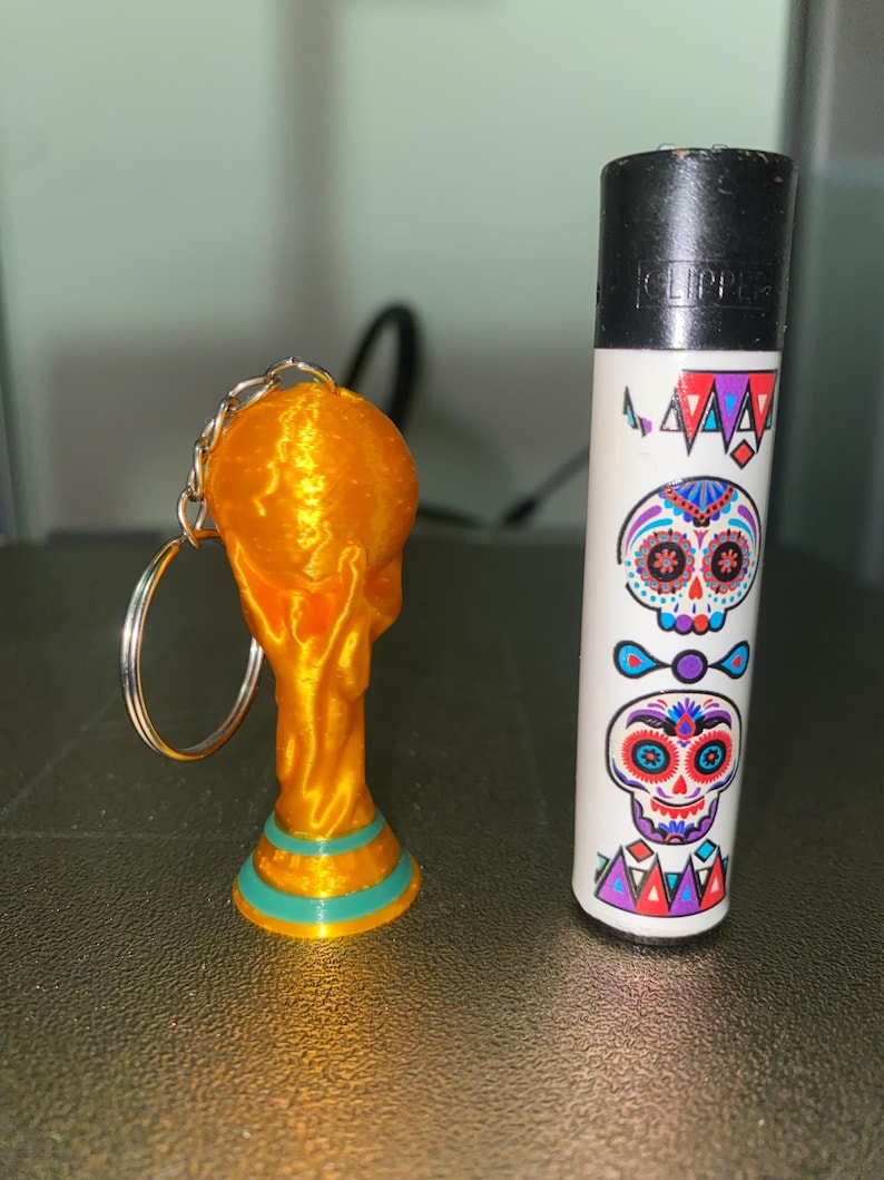 May include: A gold-colored trophy keychain next to a white lighter decorated with colorful sugar skull designs. The keychain is a miniature replica of a trophy, with a gold finish and teal accents.