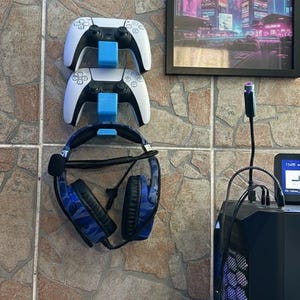 Könnte beinhalten: Eine Wandhalterung für Gaming-Zubehör mit zwei weißen und schwarzen Gamecontrollern und einem blau-getarnten Headset. Die Controller werden von blauen Halterungen gehalten. Ein gerahmtes Bild einer nächtlichen Stadt ist ebenfalls zu sehen.