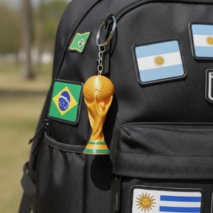 Op de afbeelding: Een goudkleurige sleutelhanger van de Wereldbeker-trofee die aan een zwarte rugzak hangt. De rugzak heeft patches van vlaggen, waaronder Brazilië, Argentinië en Uruguay. De sleutelhanger is een gedetailleerde miniatuur van de trofee.