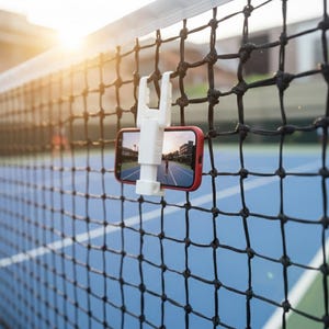 Puede incluir: Un soporte blanco para teléfono está sujeto a una red de tenis negra, sosteniendo un teléfono inteligente rojo. El teléfono muestra una escena al aire libre. La red se extiende sobre una cancha de tenis azul y verde, con luz solar en el fondo.