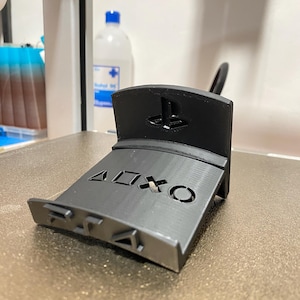 May include: A gray 3D-printed controller stand featuring the PlayStation logo and controller button symbols. The stand is designed to hold a game controller and is made of plastic.