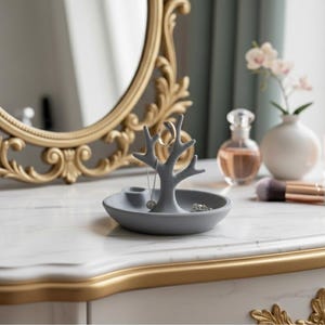 Könnte beinhalten: Ein grauer Schmuckhalter in Baumform steht auf einem weißen Marmor-Waschtisch. Ein goldgerahmter Spiegel und Parfümflaschen sind im Hintergrund. Der Schmuckhalter hat eine kleine Schale und Äste für Ringe und Halsketten.