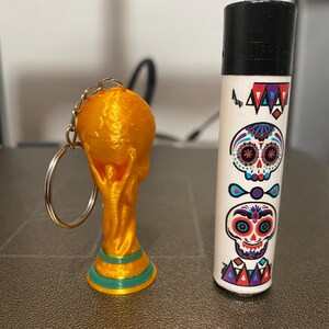 May include: A gold-colored keychain featuring a miniature World Cup trophy next to a white lighter decorated with Day of the Dead sugar skull designs. The lighter has a black top and the word "CLIPPER" printed on it.
