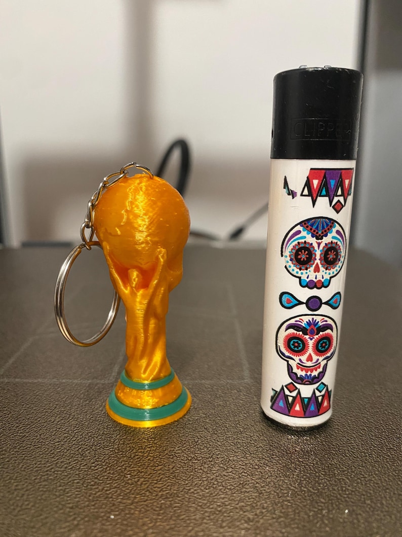 May include: A gold-colored keychain featuring a miniature World Cup trophy next to a white lighter decorated with Day of the Dead sugar skull designs. The lighter has a black top and the word "CLIPPER" printed on it.