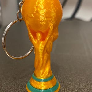 May include: A 3D-printed keychain featuring a gold-colored trophy design. The trophy has a globe held up by two figures, with a green and gold base. The keychain includes a silver chain and ring.