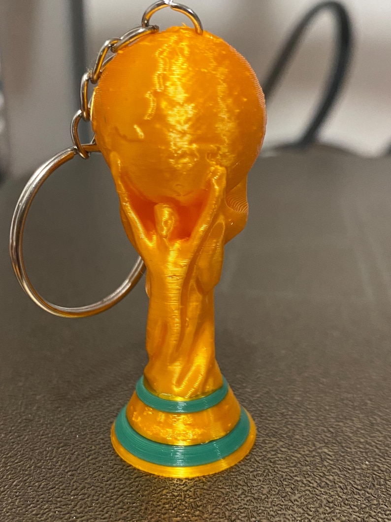 May include: A 3D-printed keychain featuring a gold-colored trophy design. The trophy has a globe held up by two figures, with a green and gold base. The keychain includes a silver chain and ring.