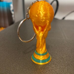 May include: A gold-colored keychain featuring a miniature trophy design. The trophy has a round top and a base with teal and gold accents. The keychain includes a silver-colored chain and ring.
