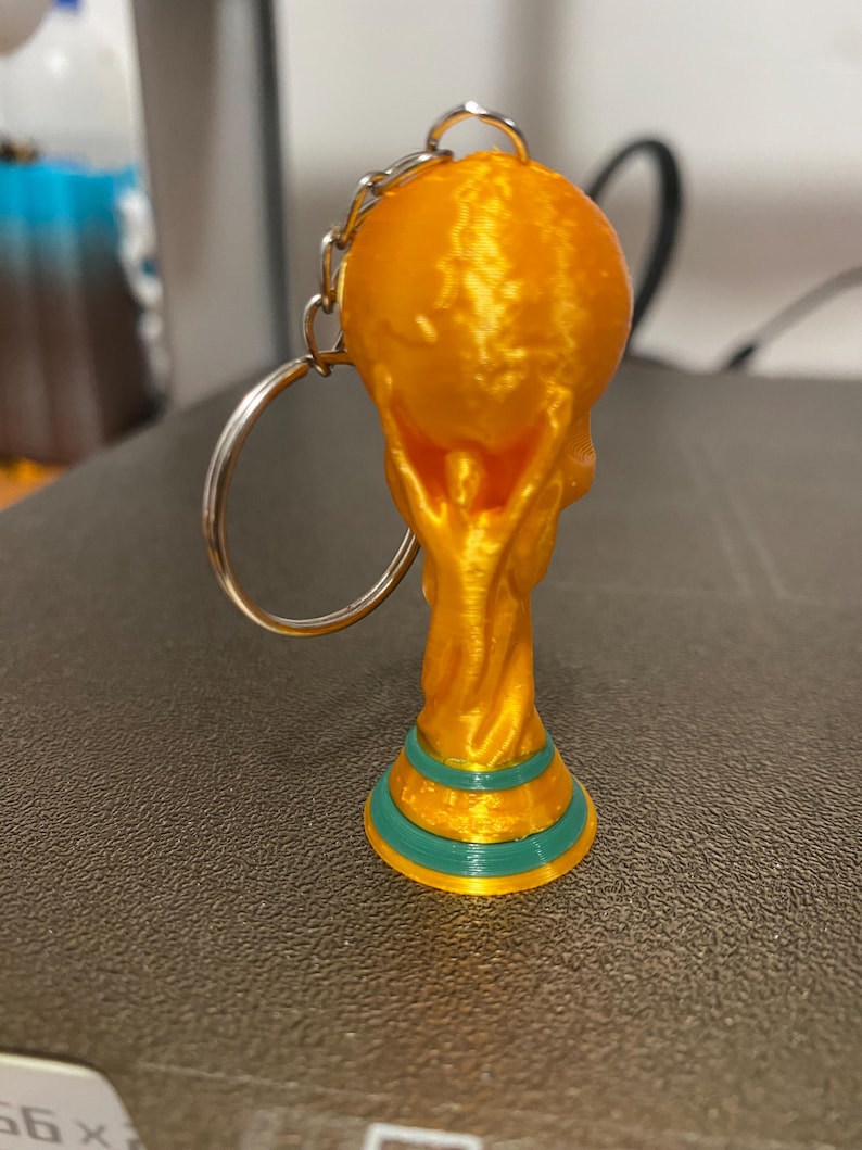 May include: A gold-colored keychain featuring a miniature trophy design. The trophy has a round top and a base with teal and gold accents. The keychain includes a silver-colored chain and ring.