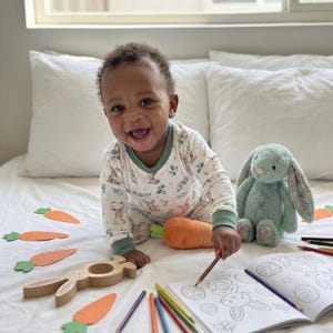 Op de afbeelding: Een lachende baby in een konijnenpyjama kleurt in een kleurboek met kleurpotloden. Een groene pluchen konijn en een oranje wortel liggen in de buurt. Houten konijn- en worteluitsteeksels zijn verspreid.