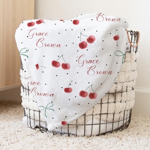Puede incluir: Una manta de felpa blanca con un estampado de cerezas y el nombre "Grace Brown" en escritura roja. La manta está doblada en una cesta de alambre con un forro color crema. Lunares negros y lazos verdes acentúan el diseño.