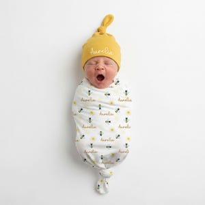 Op de afbeelding: Een pasgeboren baby gewikkeld in een witte wikkeldoek met een bijen- en madeliefpatroon, en een bijpassende mosterdgele knoopmuts. De muts en de wikkeldoek hebben beide de naam "Aurelia".