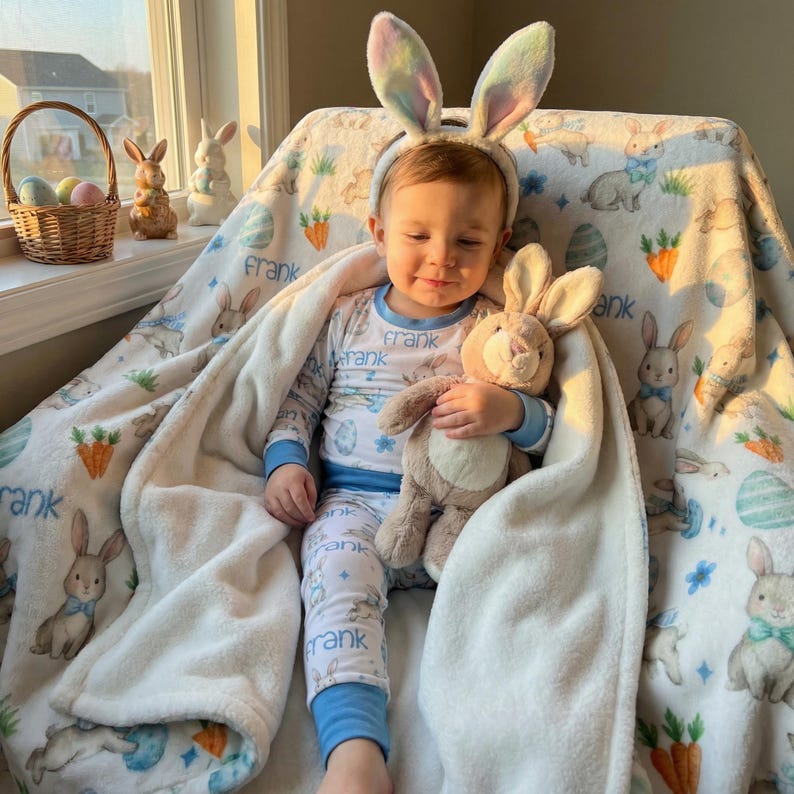 May include: A child wearing bunny ears and pajamas, holding a stuffed bunny, and wrapped in a blanket. The blanket features a bunny and carrot pattern with the name "Frank" printed on it. Easter-themed decor is visible in the background.