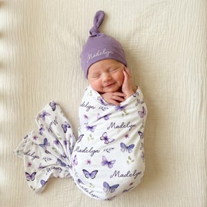 May include: A newborn baby wrapped in a white swaddle blanket with purple butterflies and the name "Madelyn" printed on it. The baby wears a matching purple knotted hat with the name "Madelyn" embroidered on it.