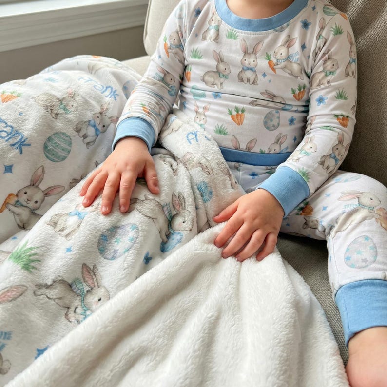 May include: White pajama set and blanket featuring a bunny and carrot pattern. The pajamas have blue trim and long sleeves. The blanket has a soft, plush texture and the name "Frank" printed on it.