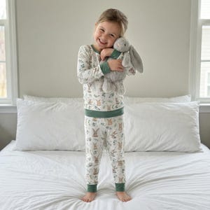 Op de afbeelding: Een kind draagt een witte pyjamaset met een konijn- en bloemenprint, met groene accenten. Het kind houdt een grijze pluchen konijn vast. De pyjama heeft lange mouwen en een broek. De achtergrond is neutraal.