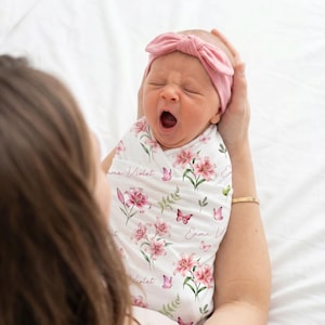 May include: A swaddled infant wearing a dusty pink knotted headband. The white swaddle blanket features a repeating pattern of pink lily flowers, green fern leaves, and small butterflies, with the name Emma Violet printed in a cursive script.