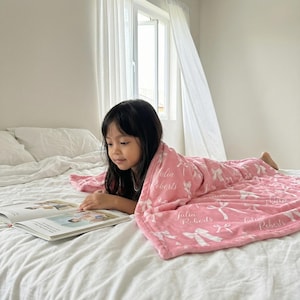 May include: A young person is lying on a bed, reading a book and covered with a pink blanket. The blanket has a white bow pattern and the name "Julia Roberts" printed on it. The room has a window with white curtains.