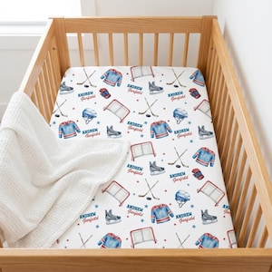 May include: A white cot sheet featuring a hockey-themed pattern. The design includes illustrations of hockey jerseys, skates, helmets, nets, gloves, hockey sticks, and stars in blue, red, and grey. The name "Andrew Garfield" is printed on the sheet.
