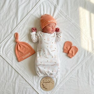 Può includere: Un neonato con un cappello, guanti e un cappello annodato abbinato color pesca, sdraiato su una coperta bianca. Il bambino indossa una tutina bianca con un motivo a coniglietti e carote. È presente anche un disco di legno con il nome "Scarlett".