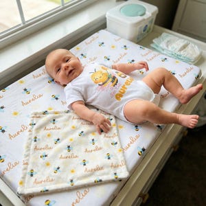 Op de afbeelding: Een baby in een witte romper met een bijenmotief en de naam "Aurelia". De baby ligt op een aankleedkussen en een bijpassend dekentje, beide met een bijen- en madeliefjespatroon. Een aankleedstation is op de achtergrond te zien.