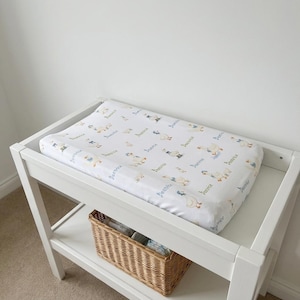 May include: A white changing pad cover featuring a repeating pattern of illustrated geese wearing blue bonnets and the name Beatrice written in a blue script font, fitted onto a rectangular changing pad on a white wooden changing table.