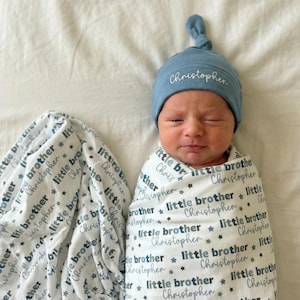 Könnte beinhalten: Ein Neugeborenes, eingewickelt in eine weiß-graue Wickeldecke und mit einer blauen Mütze. Die Decke und die Mütze sind mit dem Namen "Christopher" und den Worten "little brother" personalisiert. Die Decke hat ein Sternmuster.