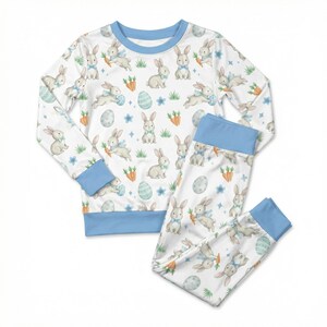 May include: White pajama set with a repeating pattern of bunnies, carrots, and Easter eggs. The long-sleeved top and pants have light blue trim. The bunny illustrations have blue bow ties.
