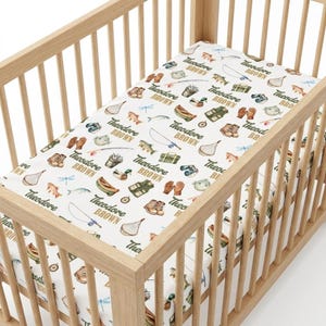 May include: A white fitted crib sheet with a repeating pattern of fishing-themed illustrations in shades of green, brown, and tan. The sheet includes fishing rods, fish, boots, and the name "Theodore Brown". The sheet is inside a wooden crib.