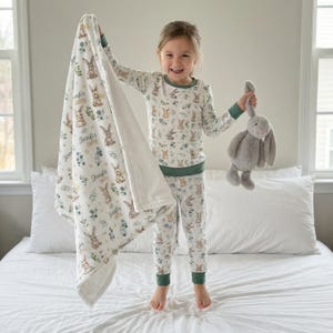 Op de afbeelding: Een kinderpyjamaset en deken met een konijnen- en bloemenpatroon. De deken is gepersonaliseerd met de naam "Jennifer Smith". De pyjama heeft groene randen. Een grijze knuffelkonijn wordt door het kind vastgehouden.
