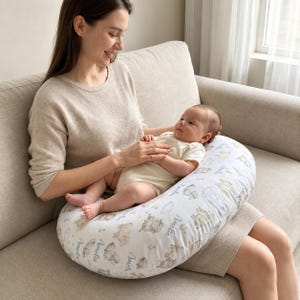 Könnte beinhalten: Ein weißes und hellgraues Stillkissen mit Schafmuster. Das kissenförmige Kissen stützt ein Baby, das von einer Person gehalten wird. Auf dem Kissen steht der Name "Amelia".