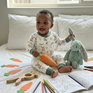 Op de afbeelding: Een lachende baby in een pyjama met konijnenprint zit op een bed met een pluchen konijn, een wortelspeeltje en een kleurboek. Oranje worteluitsparingen en kleurpotloden liggen verspreid. De pyjama heeft een witte achtergrond met konijnen- en bloempatronen.