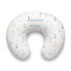 May include: A white crescent-shaped nursing pillow featuring a repeating pattern of small ducks wearing blue bonnets and green boots. The center of the pillow is personalized with the name Beatrice Winslow printed in a blue script font.