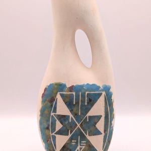 May include: A cream-coloured ceramic jug with a distinctive handle and an oval opening. The base features a blue, green, and brown design with a white geometric pattern. The jug has a smooth, curved shape.