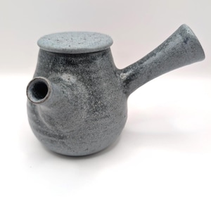 Puede incluir: Tetera de cerámica gris con tapa. La tetera tiene un pico y un asa. La cerámica tiene una textura moteada y un acabado mate. La tetera está diseñada para preparar té. Mide aproximadamente 15 cm de largo.