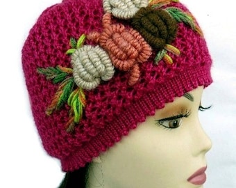 May include: A magenta knit hat with a floral design. The hat features a textured knit pattern and is adorned with three-dimensional, embroidered flowers in shades of beige, coral, and brown, with green and yellow leaves. The hat is displayed on a mannequin head.
