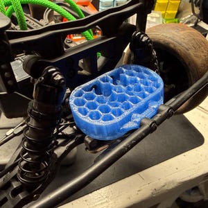 May include: A blue, honeycomb-patterned 3D-printed part is mounted on a black RC car chassis. The car has a black suspension system, a brown tire, and green wiring. A small screwdriver is visible.