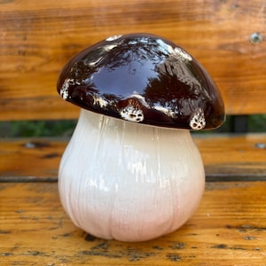 May include: A ceramic mushroom sculpture featuring a glossy, dark brown cap with small, textured white spots along the edge. The stem is a smooth, off-white color with vertical ridges, creating a realistic appearance for this decorative object.