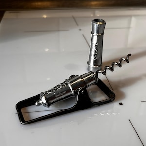 Può includere: Un cavatappi argentato a forma di cannone con un manico nero e un pezzo d'argento separato. Il cavatappi presenta un design a spirale per rimuovere i tappi dalle bottiglie. Il design evoca un tema nautico vintage.