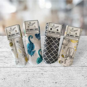 May include: Four heat-resistant pan handle covers with different patterns. The covers are made of fabric and have a label that says "Heat Resistant Pan Handle Cover". The patterns include a seahorse, a geometric design, a floral design, and a design with a mason jar.